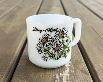 Op de afbeelding: Een witte melkglazen mok met een afbeelding van drie madeliefjes met gele harten en groene stelen. De tekst Daisy-April is in zwarte schrijfletters boven het bloemmotief gedrukt. De mok heeft een D-vormig handvat en een gladde vorm.