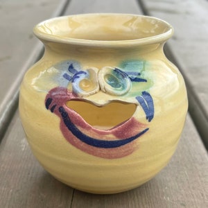 Peut inclure: Un pot en céramique jaune avec un visage peint fantaisiste. Le visage a des détails bleus, verts et roses. Le pot a une petite ouverture à l'avant.