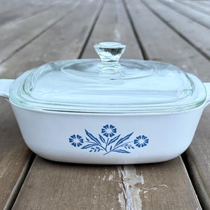 May include: White casserole dish with a clear glass lid and handle. The dish has a blue floral design. The lid has a clear glass knob. The dish is on a wooden surface.
