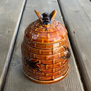 Op de afbeelding: Een bruine keramische honingpot in de vorm van een bijenkorf met een bij erop. De pot heeft een getextureerd oppervlak en is versierd met bijen.