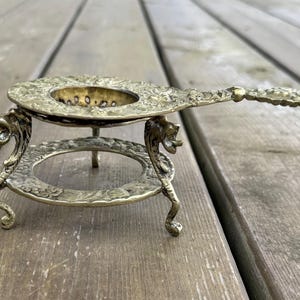 May include: A brass tea strainer with a handle and a matching stand. The strainer features an intricate floral pattern and is supported by three legs shaped like mythical creatures. The circular stand also has a matching floral embossed design.
