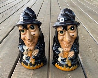 Può includere: Due statuette di streghe in ceramica. Ogni strega ha un cappello nero con fibbia argentata, occhi verdi e un lungo naso. Indossano abiti neri e tengono in mano un sacchetto di dolcetti. La base delle statuette è un calderone con bordo arancione. Decorazioni di Halloween.