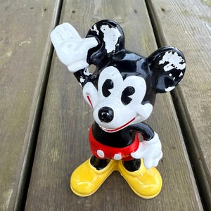 May include: A vintage Mickey Mouse figurine. The figure is black, white, red, and yellow. Mickey is waving with his right hand. The figurine has a red shorts with white polka dots, yellow shoes, and white gloves.