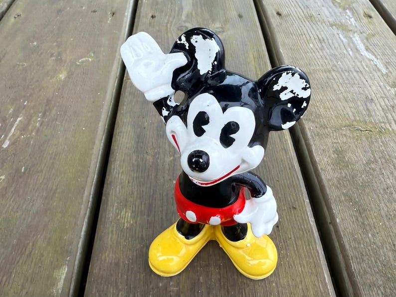 May include: A vintage Mickey Mouse figurine. The figure is black, white, red, and yellow. Mickey is waving with his right hand. The figurine has a red shorts with white polka dots, yellow shoes, and white gloves.
