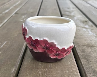 Può includere: Un vaso per piante in ceramica dal design bicolore, con una sezione superiore color crema lucido e una sezione inferiore color bordeaux scuro. Una fascia di fiori tridimensionali in rilievo, di colore rosa e bordeaux, avvolge il centro.
