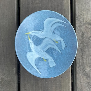 Peut inclure: Une assiette en céramique bleue avec un motif peint à la main de deux mouettes blanches en vol. Les mouettes sont peintes de manière simple et stylisée, avec des becs jaunes et des yeux noirs.