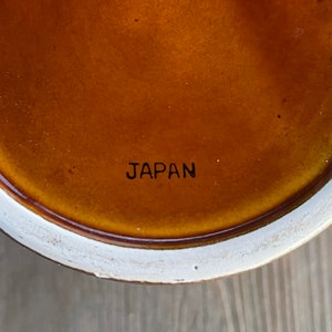 Op de afbeelding: Close-up van de onderkant van een bruin keramiek object met een witte rand. De woorden "JAPAN" zijn in zwart op de onderkant gedrukt.
