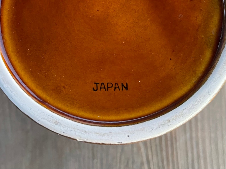 Op de afbeelding: Close-up van de onderkant van een bruin keramiek object met een witte rand. De woorden "JAPAN" zijn in zwart op de onderkant gedrukt.