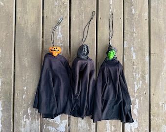 Può includere: Tre decorazioni di Halloween da appendere raffiguranti una zucca, uno scheletro e una strega, ciascuna con un mantello in tessuto nero e un cordino nero per la sospensione. Le figure hanno teste distinte e sono disposte una accanto all'altra.