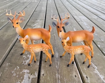Può includere: Due statuette di cervi in piedi con corna e due statuette di cerbiatti più piccoli con macchie bianche sul dorso. I cervi sono colorati in tonalità marrone chiaro, marrone e bianco, con dettagli del viso realistici e orecchie a punta.