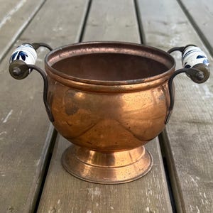 May include: A round copper bowl with a pedestal base and two curved metal handles. Each handle features a white ceramic grip decorated with a blue floral pattern. The copper surface shows a natural patina with subtle variations in colour and texture.