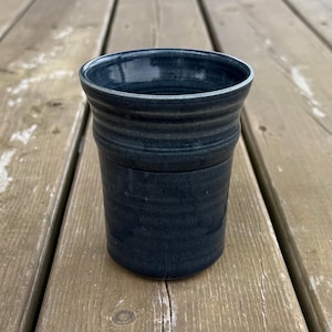 May include: A handmade, dark teal ceramic tumbler with a textured surface. The cylindrical cup has a slightly flared rim and is suitable for beverages. The pottery is displayed on a weathered wooden surface.