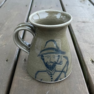 May include: A ceramic mug with a handle, featuring a hand-drawn illustration of a man with a hat, beard, and pipe. The mug is a muted green-grey colour with a blue illustration. The handle has a ribbed design.