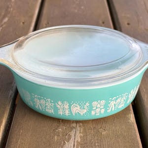 May include: A vintage turquoise Pyrex casserole dish with a clear glass lid. The dish features a white pattern of stylized figures and a rooster. The dish is on a wooden surface.