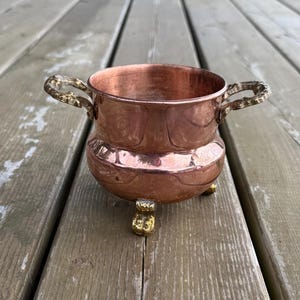 Peut inclure: Un petit bol rond en cuivre avec une finition polie, doté de deux poignées en laiton ornées sur les côtés opposés et de trois pieds décoratifs en laiton à la base. Le bol présente un sommet large et ouvert ainsi qu'un corps lisse et incurvé.