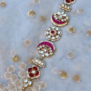May include: A gold and pink jeweled hair chain with a series of decorative pendants. The chain is made of small pearls and features a variety of shapes, including circles, crescents, and squares. The pendants are adorned with pink and white gemstones.