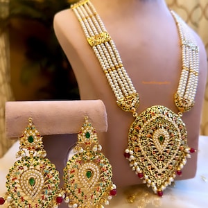 May include: A gold necklace with a large, ornate pendant and multiple strands of pearls. The pendant is decorated with green, red, and white stones. Matching earrings with similar designs are also included.