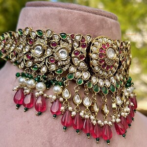 May include: A gold choker necklace with intricate floral designs, featuring white and pink gemstones, green beads, and teardrop-shaped red beads.