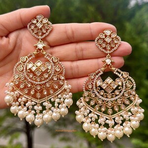 May include: A pair of gold earrings with intricate designs featuring clear stones and pearl drops. The earrings are held in a hand.