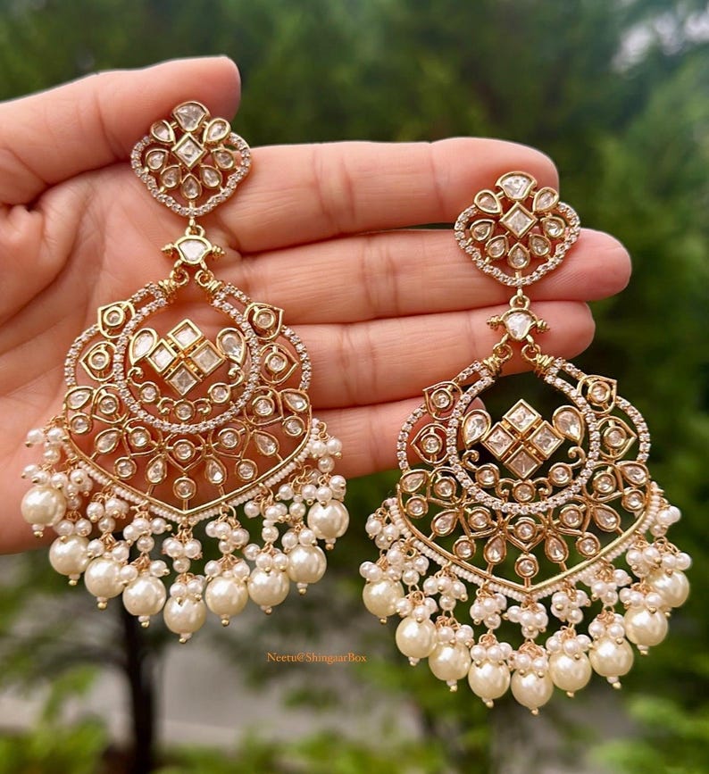 May include: A pair of gold earrings with intricate designs featuring clear stones and pearl drops. The earrings are held in a hand.