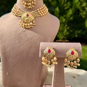 Può includere: Un set di gioielli color oro con collana, orecchini e copricapo, tutti adornati con perle finte e una gemma rossa centrale. La collana ha più fili di perle. Gli orecchini e il copricapo hanno un design abbinato.