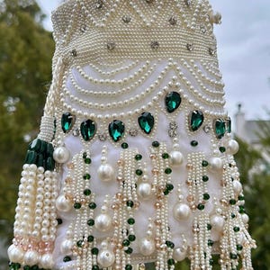 May include: White beaded handbag with pearl and emerald green details. The bag is covered in small pearls, with larger pearls and green gems. The handle is beaded, and a tassel hangs from the side.
