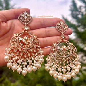 May include: A pair of gold earrings with intricate designs featuring white stones and pearls. The earrings are in the shape of a crescent moon with a heart-shaped top. The text "Neetu@ShingaarBox" is visible in the image.