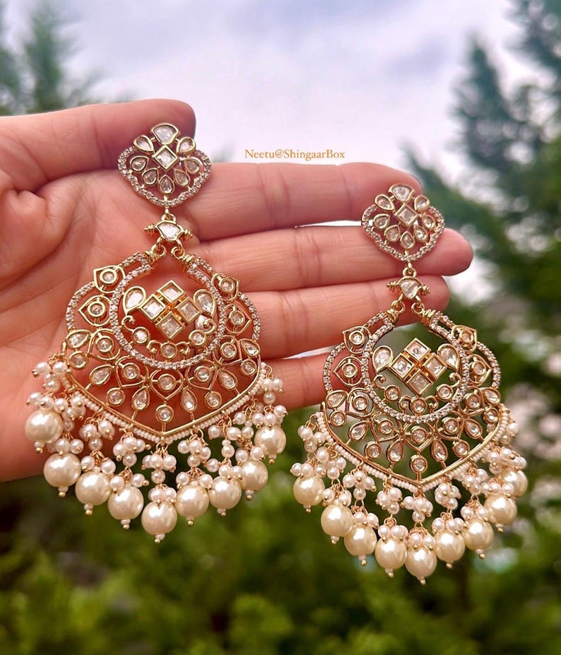 May include: A pair of gold earrings with intricate designs featuring white stones and pearls. The earrings are in the shape of a crescent moon with a heart-shaped top. The text "Neetu@ShingaarBox" is visible in the image.