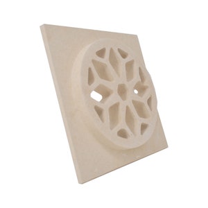 Puede incluir: Una baldosa cuadrada de cerámica de color crema con un diseño circular en relieve. El diseño es un patrón geométrico de formas entrelazadas, que se asemeja a una flor estilizada.