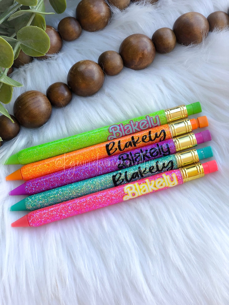 Puede incluir: Un conjunto de l&aacute;pices con purpurina en varios colores: verde, naranja, morado, turquesa y rosa. Cada l&aacute;piz lleva el nombre "Blakely" escrito en cursiva. Los l&aacute;pices tienen detalles met&aacute;licos dorados y gomas de borrar.