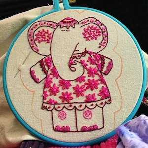 May include: Embroidered hoop art featuring a pink elephant wearing a pink floral dress. The elephant is stitched with pink thread on a white fabric background.