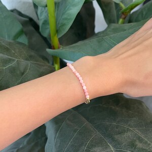 May include: A delicate pink gemstone bracelet with a gold clasp. The bracelet is made of small, round beads and is worn on a wrist.