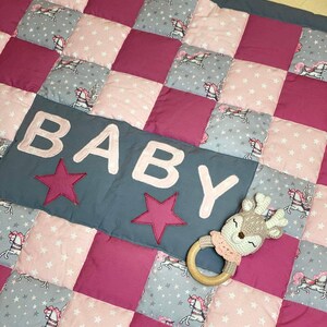 Könnte beinhalten: Eine rosa-graue Patchwork-Babydecke mit einem grauen Feld, auf dem "BABY" in rosa Buchstaben und zwei rosa Sterne stehen. Ein gehäkeltes Hirschespielzeug mit einem Holzring liegt auf der Decke.