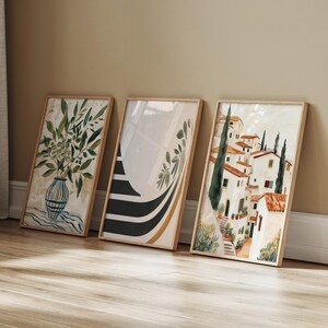 May include: Three rectangular framed wall art prints leaning against a wall. The left print shows an olive branch in a striped vase. The middle print features black and gold curved stripes with olive leaves. The right print depicts a Mediterranean village.