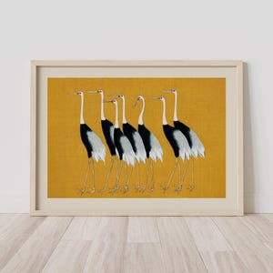 Framed art print featuring a line of stylized cranes. The birds have black and white bodies, long legs, and red eye accents. The background is a textured mustard yellow, and the frame is a light wood color. The artwork is displayed on a white wall.