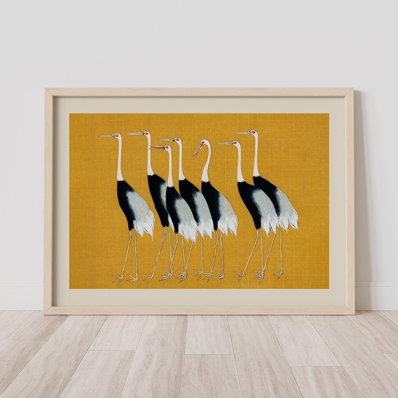 Framed art print featuring a line of stylized cranes. The birds have black and white bodies, long legs, and red eye accents. The background is a textured mustard yellow, and the frame is a light wood color. The artwork is displayed on a white wall.