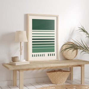 May include: A rectangular wall art print in a light wood frame featuring a cream background with horizontal stripes in varying shades of green. The text "BAUHAUS" is printed in black at the bottom left, followed by four small black vertical rectangles.