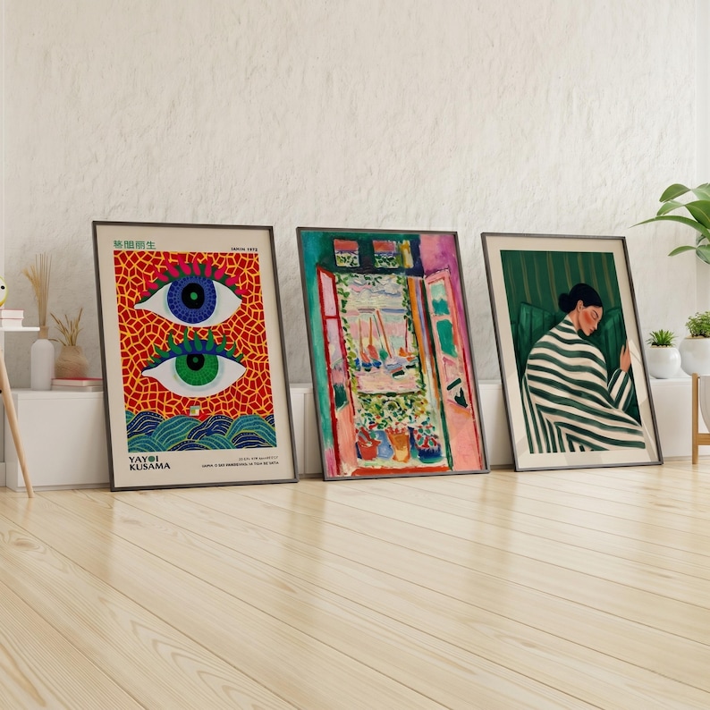Three framed art prints leaning against a white wall. The prints feature colorful abstract designs, including a pair of eyes, a window scene, and a woman in a striped robe. The frames are black, and the artwork is displayed on a wooden floor.