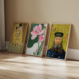 Three framed art prints leaning against a wall. The prints feature a Klimt painting, a pink lotus flower, and a portrait of a man in a blue uniform. The frames are made of light-colored wood.