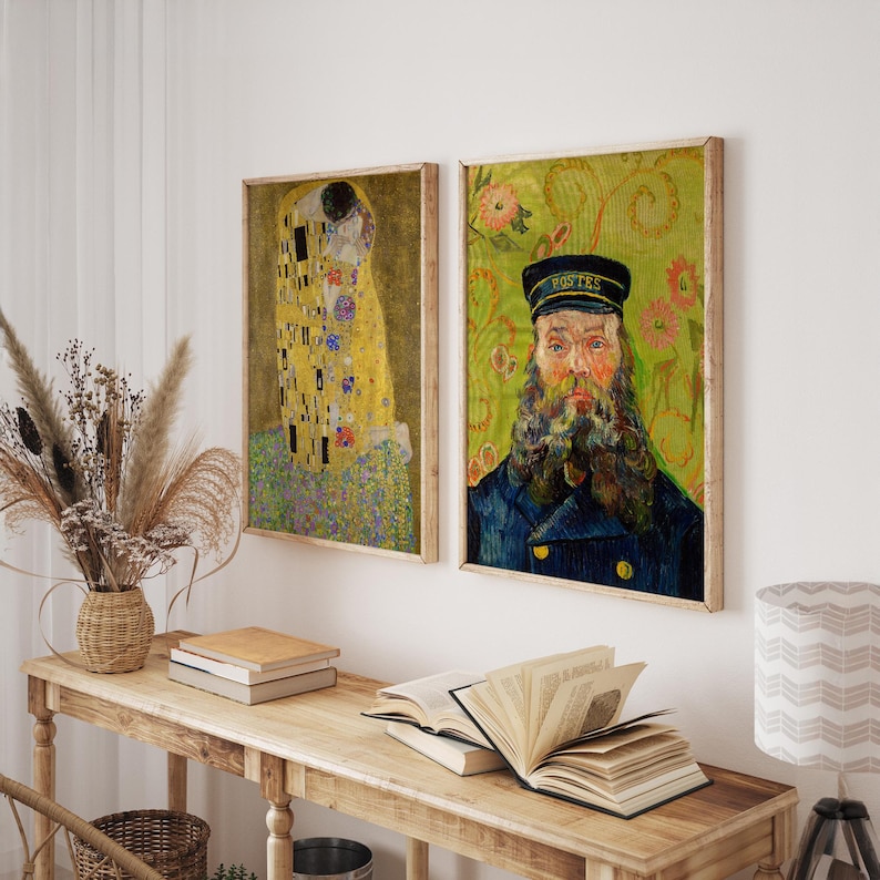 Two framed art prints hang on a white wall. One depicts a couple embracing in gold and yellow tones. The other is a portrait of a man in a blue coat and hat with the word "Postes". A wooden table holds books and a lamp.
