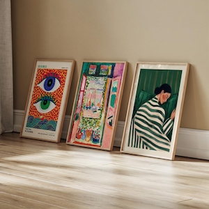 Three framed art prints leaning against a wall. The prints feature colorful artwork, including a vibrant eye design, a window scene, and a woman in a striped blanket. The frames are a light wood color.