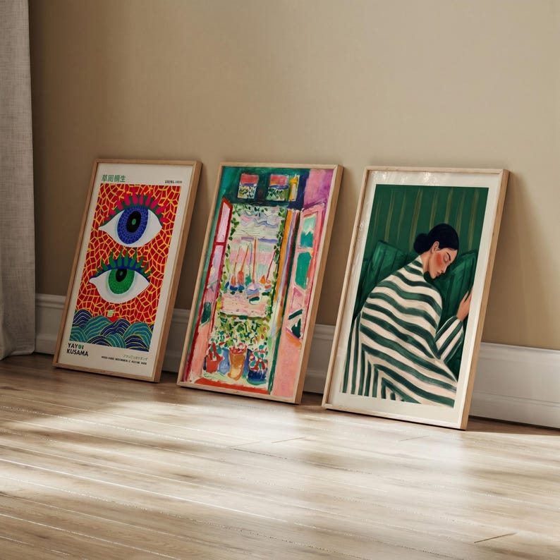 Three framed art prints leaning against a wall. The prints feature colorful artwork, including a vibrant eye design, a window scene, and a woman in a striped blanket. The frames are a light wood color.