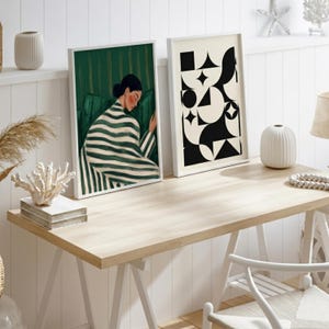 Op de afbeelding: Twee ingelijste rechthoekige kunstprints op een bureau van licht hout. De linker print toont een persoon in een groen-wit gestreept kledingstuk tegen een groene achtergrond. De rechter print toont een zwart-wit geometrisch patroon.