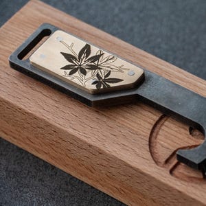 Könnte beinhalten: Ein Multitool mit Holzgriff und Metallklinge. Das Werkzeug verfügt über einen Flaschenöffner, ein Schlüsselringloch und eine dekorative Messingplatte mit Blättermuster. Das Werkzeug befindet sich in einer Holzkiste.
