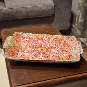 May include: Rectangular ceramic serving dish with a floral lace pattern in red, orange, and yellow hues. The dish has a scalloped edge with blue and yellow accents. The dish is placed on a dark wooden surface.