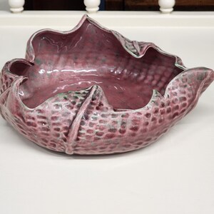 May include: A pink ceramic bowl with a textured, leaf-like design. The bowl is shaped like a flower and has a wide, open mouth.