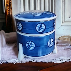 May include: A blue and white ceramic container with a lid. The container has a circular design with a blue background and white calligraphy. The container has three legs and is sitting on a white cloth.