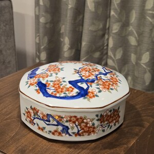 May include: White ceramic jewelry box with a lid. The box is decorated with orange flowers, blue branches, and gold trim. The box has an octagonal shape and sits on a wooden surface.