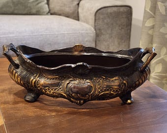May include: An antique bronze-toned decorative planter with ornate detailing. The oval-shaped planter features intricate relief patterns, handles, and small feet. The interior is a dark brown, contrasting with the gold accents on the exterior. A vintage home decor item.