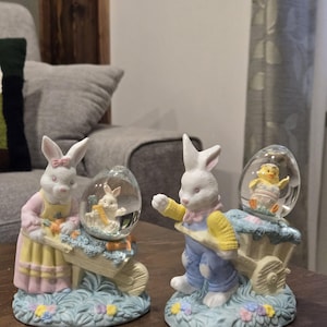 May include: Two Easter-themed figurines. One features a white bunny in a pink dress pushing a wheelbarrow with carrots and a snow globe with a bunny inside. The other has a bunny in blue overalls with a yellow jacket and a snow globe with a chick.